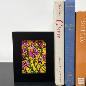 Cosmos flower painting in black frame sitting on black bookshelf with 3 books