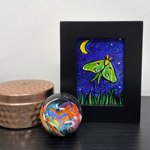 Luna Moth painting on a shelf with a trinket box and paperweight. Luna Moth painting in black wood frame features green and yellow luna moth on night sky with crescent yellow moon and white stars. Long grasses dance along the bottom of the scene.