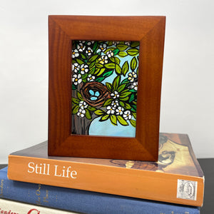 Robin&#39;s Nest painting sitting on top of books. Painting shows blue robin&#39;s eggs in a brown nest in a tree with flowers and leaves. Light blue sky is visible between the branches.