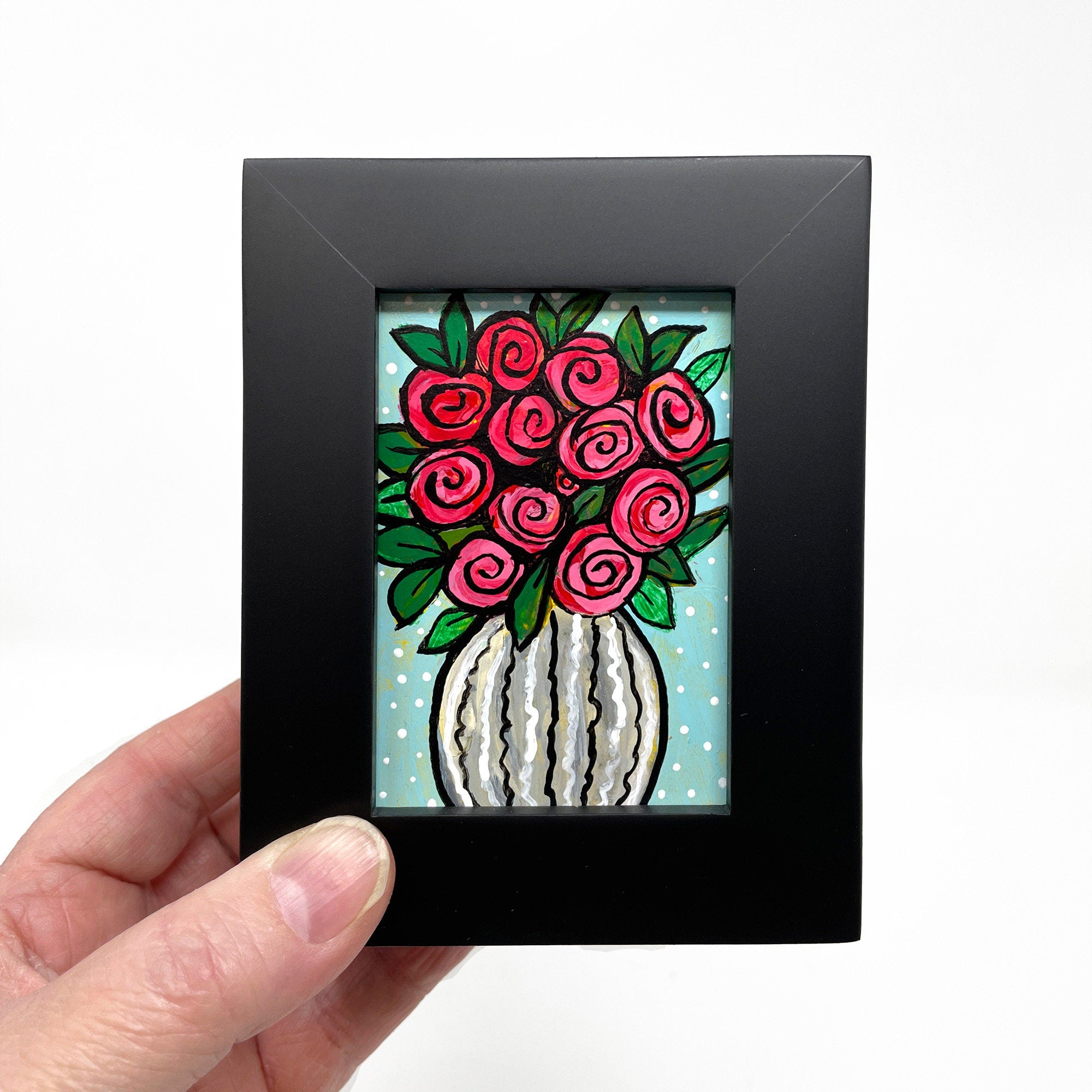 Hand holding Vase of Roses painting in black frame - White striped vase with red roses and green leaves. Light blue background with white polka dots.