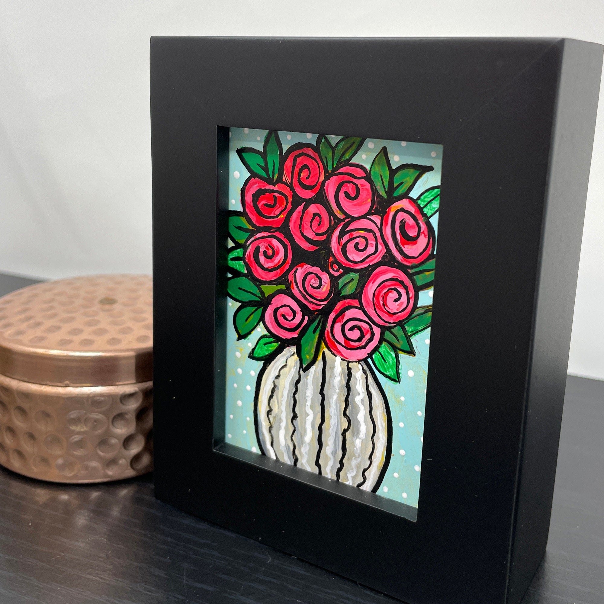 Side view of Vase of Roses in black painting sitting on black table with copper box.