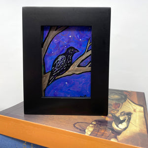 Small raven painting in black frame sitting on top of books